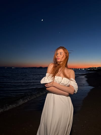 A woman with long, reddish-brown hair stands on a dark, pebbly beach at dusk. She is wearing a white off-the-shoulder dress with puffed sleeves and her arms are crossed. She is looking to her left, out at the sea. The water is dark and the waves are gently lapping at the shore. In the distance, along the coastline, there are lights from a city or town. The sky is a deep blue transitioning to orange and pink near the horizon. A crescent moon and a bright star are visible in the upper portion of the sky. The location is the coast of Paleo Faliro, Greece.