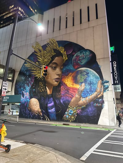 A vibrant mural on a building's side wall features a woman's portrait, adorned with a gold floral headpiece. She lies against a backdrop of swirling, cosmic imagery that includes planets and a hand holding a glowing globe. A traffic light partially obscures the mural. Below, street signs and a garage entrance are visible, along with portions of the sidewalk and street. The upper part of the building displays a sign that reads "ONE SOUTH DISTRICT ONE".