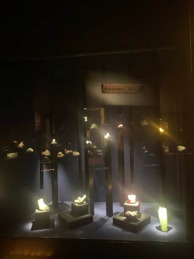 The image depicts a display case of minerals, likely in a museum or exhibit in Grenoble, France, as indicated by the visible text on a sign "Musée Grenoble". The objects are lit from above, highlighting the various rock and mineral samples. The display consists of vertical black bars with platforms holding the minerals. Some minerals are translucent and illuminated by backlighting. The overall scene is dimly lit, focusing the attention on the displayed items.