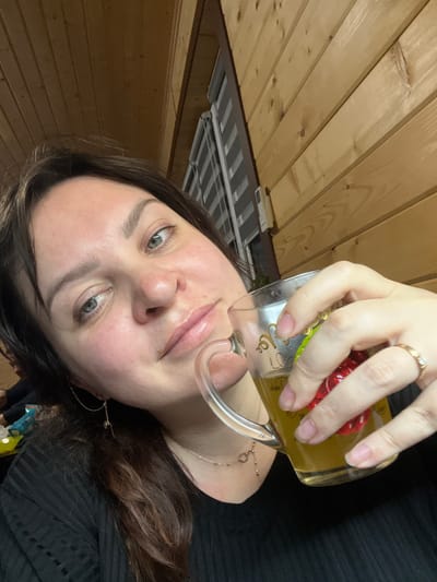 A woman with brown hair and light eyes is holding a glass mug filled with a yellow liquid. She is wearing a ring on her left hand. The background includes a wood-paneled wall and a ceiling fixture. The woman is wearing a dark-colored top and a gold chain. There is no information to suggest that the location is Bol'shakino, Russia.