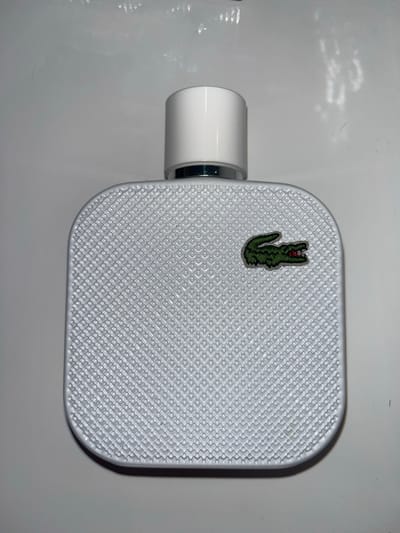 A white, rectangular object, likely a perfume bottle, is presented from a top-down view, resting on a smooth, light grey surface. The main body of the object is white and features a distinctive textured pattern of small, embossed squares or interlocking 'L' shapes, arranged diagonally across its entire surface.
Near the upper left corner of the object, a small, embroidered green crocodile logo with subtle red highlights is affixed, clearly identifying it as the Lacoste brand emblem. A white, cylindrical cap with a reflective silver band at its base extends from the left side of the rectangular body.
The background is an uncluttered, light grey expanse, possibly a table or countertop, which exhibits some faint, soft reflections and minor smudges, particularly in the upper and lower left sections. The lighting is uniform, suggesting an indoor environment. No human subjects are present, and no activity is taking place; the object is static. There is no visible text apart from the brand logo. While the location context is Laval, Canada, there are no specific visual cues within the image to indicate this.