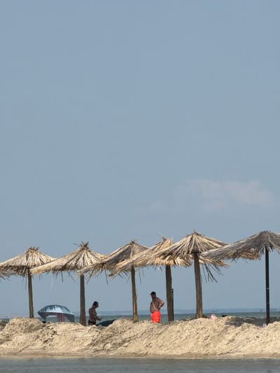 The image depicts a beach scene with a clear blue sky overhead. In the foreground, a sandy shoreline is visible, with several thatched umbrellas providing shade. Several people are scattered around the beach. One man in orange shorts stands near an umbrella, looking away from the camera. Another person, partially obscured, is seen near a blue umbrella. The water is calm and extends to the horizon, where a thin line of land is visible. The overall atmosphere is one of a peaceful, sunny day at the beach.