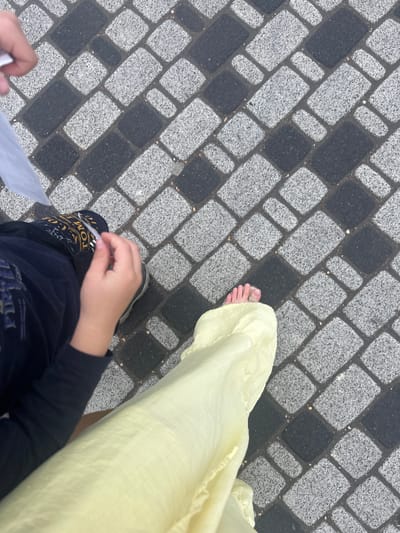 The image shows a first-person perspective of someone walking on a paved pathway composed of rectangular stones in a black and grey checkerboard pattern. To the right, the lower part of a person's leg is visible, wearing a light yellow fabric that appears to be a long skirt or dress, and their bare foot. To the left and lower in the frame, another person's hands and legs are visible. This person is wearing dark clothing, including a long-sleeved shirt and pants with visible gold text. One hand is holding a piece of white paper. The scene appears to be outdoors, possibly in a park or pedestrian area. The lighting suggests it could be daytime, and no specific weather conditions are evident. The presence of two people, one walking and the other partially visible, suggests they are together, perhaps strolling or pausing. The visible text on the pants includes the words "TOM" and other stylized lettering in gold. The context provided states the location is Novoselye, Russia.