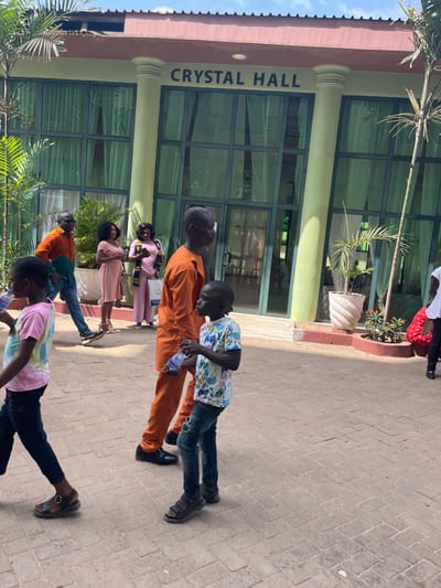 The image displays an outdoor scene in Madina, Ghana, in front of the "CRYSTAL HALL" building. The building features a light-colored facade with two prominent light green columns and a reddish roof. Its main entrance consists of multiple dark-framed glass panels, behind which light green curtains are visible. The name "CRYSTAL HALL" is displayed in dark capital letters above the entrance.
The ground is paved with reddish-brown interlocking bricks. Lush green foliage, including palm trees, is visible on both sides of the frame and in pots near the building. The scene is brightly lit by sunlight, indicating clear weather and casting visible shadows.
Seven individuals are present. In the foreground, a young child, likely a boy, wearing a pink and white tie-dye t-shirt and dark trousers, walks towards the left. Behind him, an adult male in an orange, blue, and green color-block shirt and blue jeans walks left. Further back, two adult females are standing; one wears a light pink sleeveless dress and holds a handbag, while the other wears a pink jumpsuit with a patterned stole and carries a white bag. In the mid-ground, an adult male in a long-sleeved orange tunic walks towards the right, holding a clear plastic water bottle. Beside him, a young child, likely a boy, wearing a colorful patterned t-shirt, distressed blue jeans, and sandals, also walks right, holding a clear plastic water bottle. A seventh individual is partially visible on the far right side of the frame.