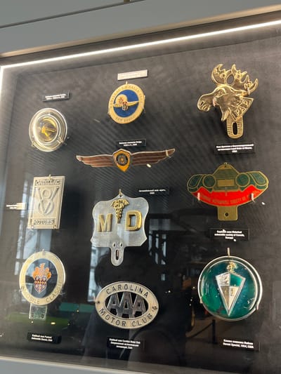 This media file showcases a collection of vintage automobile badges and emblems displayed in a glass case. The objects are arranged in rows on a dark background. Visible text on the labels indicates they are from various automobile clubs and historical societies, including "Cadillac La Salle Club San Diego," "Historical Automobile Society of Canada," and "Carolina Motor Club." Some of the emblems themselves have visible text such as "CADILLAC," "MOTORS," and "CLASSICS." There are also specific vehicle models mentioned like "Hudson Hornet Special." The lighting suggests an indoor setting, likely a museum or exhibition, with bright lights illuminating the display. The collection appears to be a historical exhibit dedicated to automotive memorabilia. The location context provided is Verkhnyaya Pyshma, Russia, which might be the location of this display. No people are visible in the media file, and the focus is entirely on the objects.