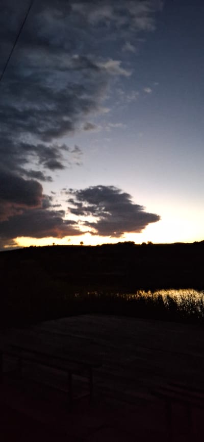 The image depicts a twilight scene with a dramatic sky over a rural landscape. The sky is transitioning from a deep blue at the top to lighter shades of purple and orange near the horizon. Dark, textured clouds are scattered across the upper portion, with some illuminated by the setting sun. A thin, faint line, possibly a power line, runs diagonally from the upper left corner.
Below the sky, a silhouetted landscape features a dark, undulating horizon line. Two small, solitary trees stand out against the bright band of light on the horizon. In the midground, a body of water reflects the warm glow of the sunset, with reeds or tall grass visible along its edges. The foreground is dominated by the dark silhouette of a wooden structure, possibly a bench or table, with visible legs and a textured surface.
The location is identified as the vicinity of Ablanitsa, Bulgaria. The scene evokes a sense of calm and natural beauty characteristic of a rural sunset.