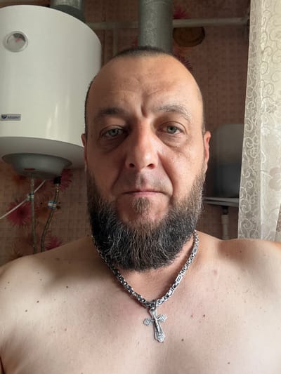 A man with a beard and a silver cross necklace is pictured from the chest up, in what appears to be a bathroom in Zhirnov, Russia. He is looking directly at the camera with a neutral expression. Behind him, a white water heater is visible on the left, with pipes and floral wallpaper. A sheer white curtain hangs on the right. The man's skin is fair, with visible pores and some moles. His beard is dark and neatly trimmed. The overall lighting is bright, suggesting it might be daytime.