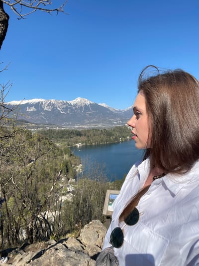 A woman stands on an elevated rocky area overlooking Lake Bled and its surroundings in Slovenia. The lake is in the background, with a mountain range visible on the left. The sky is clear and blue. The woman has long brown hair and wears a white shirt with sunglasses hanging from it. The immediate foreground consists of rocks and bare branches.