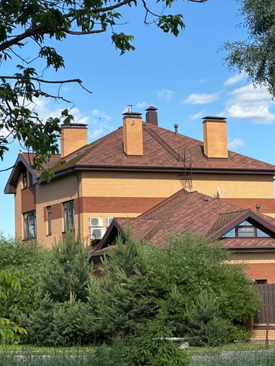The image shows a two-story house with a reddish-brown asphalt shingle roof. The house is constructed with two-tone brickwork, featuring bands of yellow and darker red bricks. Several chimneys with metal caps are visible on the roof. Two air conditioning units are mounted on the exterior wall of the house. The house is surrounded by lush green trees and bushes. In the background, a clear blue sky with scattered white clouds is visible. The scene is located in Dubrovitsy, Russia.
