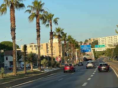 The image shows a road in Toulon, France, lined with palm trees. There are several cars driving on the road, including a red SUV, a silver SUV, and a black sedan. There are also several buildings visible in the background, including a tall, modern apartment building and a more traditional building. The sky is clear and blue, and the sun is shining. There are also a few traffic signs visible, indicating directions to Nice, Hyeres, Toulon, Sainte Musse, Cap Brun, and La Rode. One sign indicates a 4.3-meter height restriction. The overall scene suggests a sunny day in a coastal city.