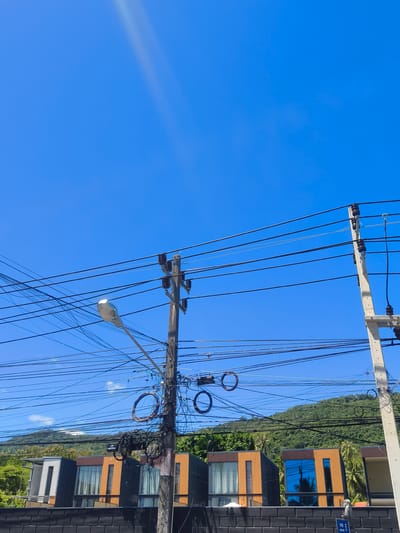 This is a full shot, eye-level, outdoor, daytime, vertical photograph of a street scene in Baan Thong Po, Thailand. The clear, bright blue sky dominates the upper portion of the image. In the foreground, a cluster of utility poles with numerous tangled black electrical wires spans across the frame, creating a dense web of lines against the sky. One pole features a street lamp. The poles are concrete with some wooden elements.
Below the wires, modern, multi-story buildings with dark grey and orange facades are visible. Some of these buildings have large windows, some of which reflect the blue sky and palm trees. A dark, textured wall runs along the bottom of the frame in front of the buildings. In the background, a lush, green, tree-covered hill rises under the blue sky. The overall impression is of a tropical urban environment with a heavy concentration of overhead electrical infrastructure. There are no people or animals visible in the image.