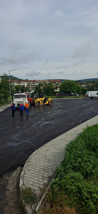 The image depicts a street paving operation in the city of Strazhitsa, Bulgaria. A white truck and a yellow road roller are positioned on a freshly paved asphalt surface, which is still wet and appears to be a parking lot or an intersection. Several individuals in work attire are present, seemingly overseeing or participating in the paving process. A light pole stands to the left, and residential buildings are visible in the background. The sky is overcast, suggesting cloudy weather. The foreground features a paved walkway and dense green vegetation.