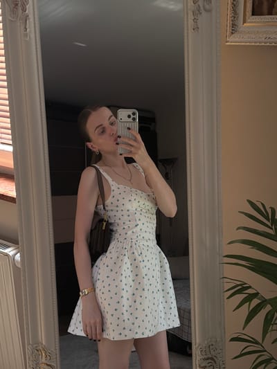 A woman is taking a mirror selfie in what appears to be a bedroom. She is wearing a white dress with a delicate blue floral pattern and a thin gold necklace. A brown crossbody bag is slung over her shoulder, and she wears a gold watch on her left wrist. Her right hand is holding a silver smartphone, taking the picture. She has dark nail polish on. The mirror she is using is large and ornate, with a decorative white frame. In the reflection, a corner of a framed painting can be seen on the wall. A large leafy plant is also visible in the reflection, suggesting the presence of natural light, possibly from a window. The overall impression is casual and domestic. The image is oriented vertically, and the woman's reflection appears to be sideways, indicating the photo was taken with the phone held horizontally. The lighting is soft, suggesting it might be daytime, but it is difficult to determine the exact time of day or weather conditions. There is no visible text in the image.