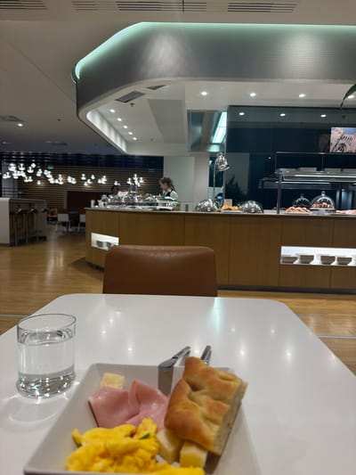 The image captures an indoor scene, likely a modern dining area or lounge, with a focus on a breakfast or brunch setting. The perspective is from a slightly elevated angle, looking across a table towards a buffet area in the background.
In the immediate foreground, on the right side, a pristine white table holds a clear glass of water. A white rectangular plate is laden with a meal, consisting of several pieces of light-colored, airy bread (resembling focaccia), neatly folded slices of pink ham, bright yellow scrambled eggs, and small cubes of pale yellow cheese. Silver cutlery is partially visible beside the plate. A brown, leather-upholstered chair is positioned to the left of the table, suggesting a diner's seat.
The mid-ground reveals a contemporary buffet or serving counter. It features a long wooden base with a darker, reflective top. Numerous silver cloches cover various food items, and pastries are arranged on trays. A person with dark hair, wearing a white shirt, stands behind this counter, seemingly attending to the buffet.
The background showcases the modern architecture of the space. A distinctive, curved ceiling feature with a perforated texture and subtle green lighting runs along the left side. Recessed spotlights illuminate the white ceiling. The flooring is polished wood. Further back, a cluster of modern, spherical hanging light fixtures adds to the ambiance. A few individuals are visible in the distance, some seated at tables. On a wall in the far upper right, an image of an architectural structure, possibly a cathedral or historical building, can be seen.
The setting suggests a breakfast or brunch service in a clean, contemporary environment, possibly an airport lounge or a hotel dining room, fitting the context of Ferno, Italy (home to Milan Malpensa Airport). The artificial lighting indicates an indoor setting, with no specific time of day discernible from natural light. No explicit text is visible in the image.