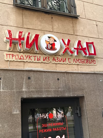 The image shows the exterior of a store named "НИ ХАО" (Ni Hao) in Oktyabrsky, Russia. The store's name is displayed in large red letters with white outlines, above a banner that reads "ПРОДУКТЫ ИЗ АЗИИ С ЛЮБОВЬЮ" (Products from Asia with Love). A cartoon image of a smiling Asian man holding a bowl of food is part of the signage. Below the main sign, on the glass door of the store, the name "НИ ХАО" is repeated in smaller red letters, along with "ЭШ РЕЖИМЫ" (Work Schedules) and "РЕЖИМ РАБОТЫ" (Work Hours). The building facade is made of light-colored stone, and a barred window is visible above the sign. The entrance is a dark-colored door with reflections of trees and buildings.