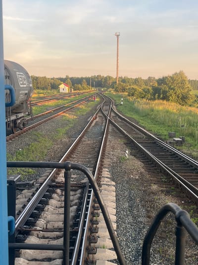 The image depicts a railway yard in Kauchuk Stantsiya, Russia, under a clear sky with a warm, golden light suggesting either sunrise or sunset. The foreground shows the perspective from a train, with a blue metal railing and part of a dark-colored train car on the left. Multiple sets of train tracks stretch into the distance, some with grass growing between the gravel. In the mid-ground, a small white building with a red roof is visible on the left side of the tracks. Further into the yard, a tall, slender utility pole stands prominently on the right. The tracks diverge and converge, indicating a junction point. The surrounding environment is characterized by dense green trees and vegetation.