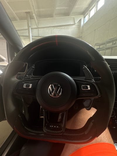 The image shows a close-up view of a car's steering wheel, which appears to be modified with carbon fiber accents and red stitching. The Volkswagen logo is visible in the center of the steering wheel. There are also paddle shifters behind the wheel. The background suggests the interior of a garage or workshop, with exposed pipes and fluorescent lighting visible on the ceiling. The lower part of the image shows a person's legs, wearing shorts, with one leg slightly blurred. There is no indication of the specific location in Schkopau, Germany.