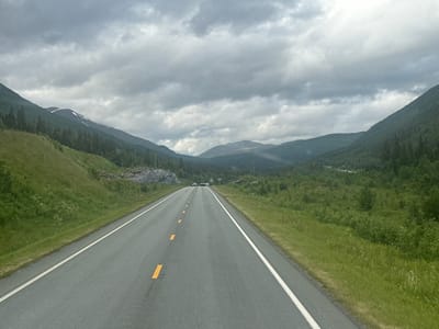 A wide asphalt road with white and yellow lane markings stretches into the distance, flanked by grassy verges. The road disappears into a valley between tree-covered mountains under a cloudy, overcast sky. The mountains on either side are steep and covered in a mix of dense green foliage and some patches of snow or lighter rock on the higher elevations. The overall scene conveys a sense of travel through a vast, natural landscape. There are a few vehicles visible on the road in the distance, suggesting it is an active route. The time of day appears to be daytime, given the brightness, but the heavy cloud cover creates a subdued, possibly cool atmosphere. The location cues, such as the mountainous terrain and the type of road, are consistent with a remote or rural setting. The weather is clearly overcast, with the clouds filling much of the upper frame, casting a diffused light over the scene. No text is visible in the image.