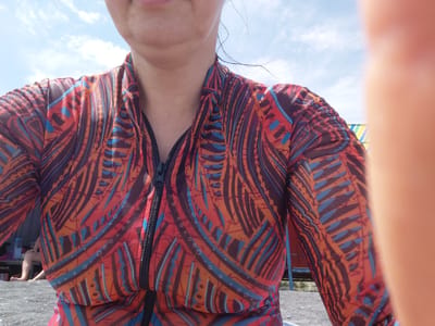 A close-up, head-on, medium shot captures a woman from the collarbones to the waist, wearing a long-sleeved, zip-front rashguard with a vibrant, abstract pattern of orange, blue, and dark red. Her mouth is visible at the top of the frame, and her neck, with a visible water droplet, is also in view. The background suggests an outdoor, possibly coastal or lakeside location, with a bright sky and wispy clouds above. In the lower left corner, a dark, gravelly surface is visible, hinting at a beach or similar terrain. Further back, a person's legs are partially visible, seated near a table with a bottle of water. A colorful, striped structure, possibly part of a beach umbrella or shelter, is visible in the background to the right. The image appears to be taken in Ақши, Kazakhstan.