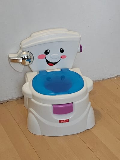 A Fisher-Price potty training toilet is pictured indoors, positioned on a wooden floor against a light-colored wall. The potty is white with a blue seat and a pink button on the front. It has a smiling face design on the front with large black eyes and pink cheeks. A chrome handle is attached to the left side of the toilet. The environment does not provide any specific details to suggest a location within Trier, Germany.