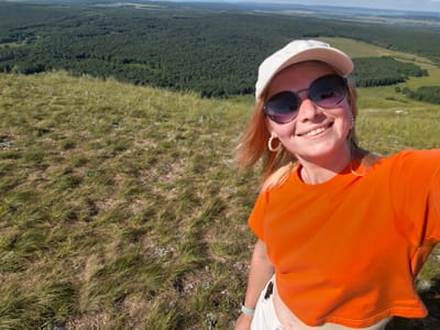 A young woman with blonde hair, wearing a white cap and sunglasses, takes a selfie on a grassy hill overlooking a vast, wooded valley in the city of Shikhan, Russia. She is dressed in an orange crop top and white shorts. The landscape is dominated by rolling hills covered in dense green forest and a bright, sunny sky. The foreground shows dry, yellowed grass typical of a hilltop environment. The woman is smiling and her face is well-lit by the sun.