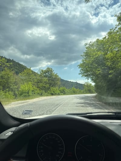 The image provides a driver's or passenger's perspective from inside a vehicle, looking out at a scenic road and natural landscape. The setting is likely rural or mountainous, consistent with Cherna Mesta, Bulgaria.
In the foreground, the dark grey/black dashboard and steering wheel of the car are visible on the right. Part of the instrument cluster is in view, displaying a speedometer marked up to 240 km/h and a tachometer reading up to 7 x 1000 r/min. Small red indicators are also present on the gauges. A black windshield wiper blade is positioned on the left side of the windshield. A light-colored sticker with unclear markings is affixed to the bottom-left of the windshield.
Beyond the vehicle's interior, a paved road extends into the distance, with shadows from overhead trees dappling its surface. The road is flanked by abundant, vibrant green trees and bushes, which appear to be in full leaf, suggesting a spring or summer season. In the midground, verdant hills or mountains rise, covered in dense forestation.
The sky above is partly cloudy, featuring large grey and white clouds that partially obscure the sun, though patches of blue sky are visible. The bright, diffused lighting indicates it is daytime, likely during the late morning or early afternoon. The overall scene depicts a calm journey through a picturesque, natural environment.