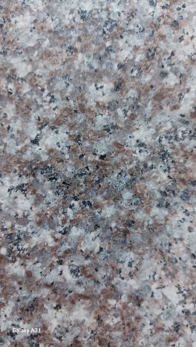 The image is a close-up, textured, abstract view of a speckled granite surface. The granite displays a pattern of irregular flecks of white, light grey, brown, and black. The lighting appears to be ambient, casting no harsh shadows and creating a generally flat and evenly illuminated surface. No people, objects, or actions are discernible. The watermark "Galaxy A71" is visible in the bottom left corner of the original image. No elements in the image relate to the city of Umm Salal Ali, Qatar.