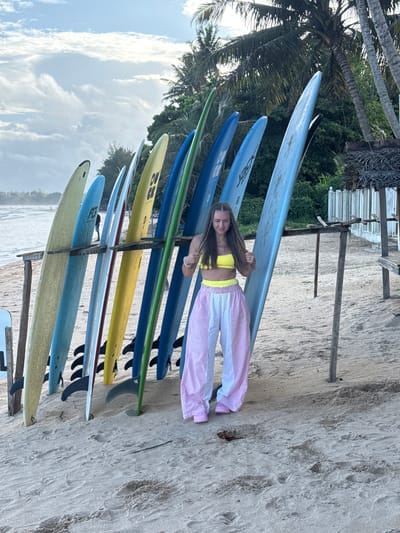 A young woman with long braided hair, wearing a yellow bikini top and pink and white striped pants, leans against a row of surfboards on a sandy beach. The surfboards are propped up on a wooden rack, with several different colors visible including blue, yellow, and green. Palm trees and lush green foliage are in the background, suggesting a tropical location. The sky is bright and cloudy, indicating it's daytime, likely with good weather for surfing. The woman appears relaxed and content, with a slight smile. The scene is set in Weligama, Sri Lanka, a well-known surfing destination. Some text is visible on one of the surfboards, possibly a brand name or a number, but it is not clearly legible. The overall impression is one of a leisurely day at the beach, enjoying the surf culture.