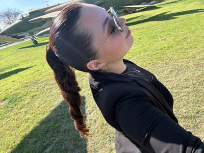 A woman with dark, shiny hair in a high ponytail gazes upward, wearing sunglasses. She's dressed in a black blazer over a dark top. She has gold earrings. Behind her is a grassy field, with a military aircraft partially visible. The sun seems bright, casting shadows across the lawn. The photo appears to be taken outdoors, possibly at a military museum or park.