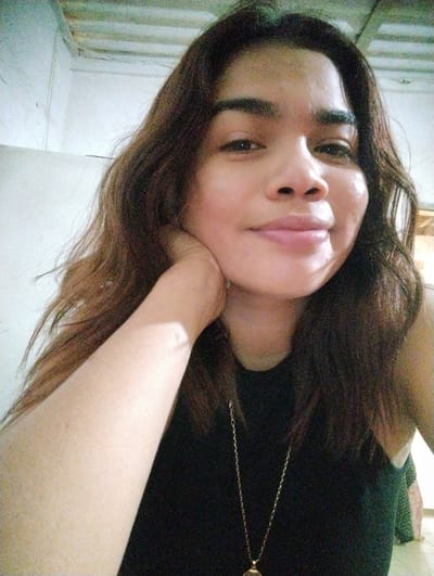A young woman of indeterminate age and ethnicity is depicted in a close-up, selfie-style photograph. She has dark brown hair that falls past her shoulders and frames her face. Her right arm is raised, and her hand rests on her chin, supporting her head as she looks directly at the camera with a slight smile. She is wearing a black sleeveless top and a gold chain necklace with a pendant. The background is out of focus but appears to be an indoor setting with a white wall and a ceiling that suggests a rustic or unfinished structure. The lighting suggests an artificial light source, possibly a flash, as evidenced by highlights on her face and arm. The image quality is moderate, with some graininess. There is no visual information to indicate the location in Punta de Piedras, Venezuela.