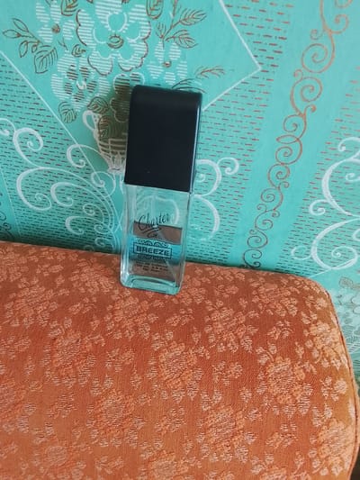 The image shows a close-up shot of a bottle of Charter Breeze eau de toilette for men. The bottle is rectangular with a black cap and a light blue liquid inside. The label on the bottle is white with blue text. The bottle is sitting on a textured orange surface, possibly a sofa or a piece of upholstery. In the background, there is a teal-colored fabric with a floral pattern in white and gold. The lighting suggests an indoor setting. There is no indication of the city of Bender, Moldova, or any other specific geographical location within the image.