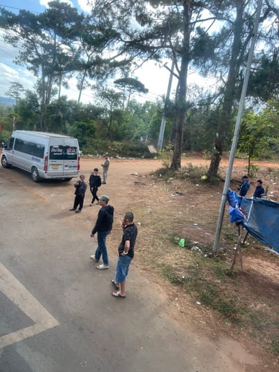 A group of people stand near a white van on a road. One man wears a black shirt and jeans, another a black jacket. Others stand closer to the van, which has lettering on the back. A blue tarp is suspended overhead in the background, with more people nearby. The scene is surrounded by trees and foliage, with trash scattered on the ground. The photograph is taken from an elevated position, slightly tilted.