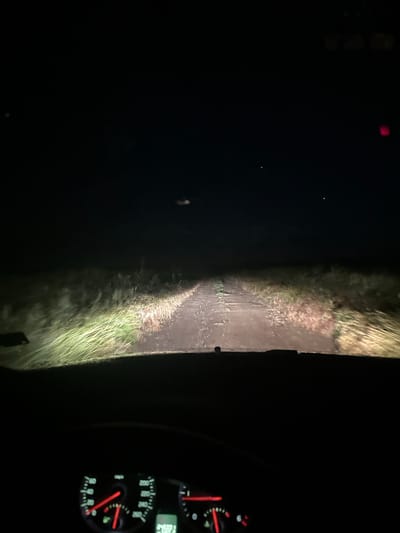 The image shows a view from inside a car driving down a dark road in Jitnitsa, Bulgaria at night. The road appears to be unpaved, with vegetation along both sides. The car's headlights illuminate the road ahead. The car's dashboard and instrument panel are visible on the right side of the image, showing the speedometer and other gauges. The image is dark with limited visibility.