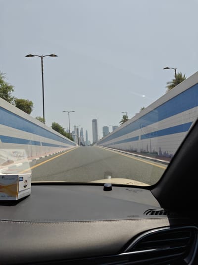 The image is taken from inside a vehicle, looking forward through the windshield at a road in Dubai, United Arab Emirates. The road is bordered by elevated walls adorned with alternating blue and white horizontal stripes. In the distance, several modern skyscrapers are visible against a clear, bright sky. A box of tissues sits on the dashboard in the foreground. The image captures a typical urban landscape in Dubai, characterized by its futuristic architecture and well-maintained infrastructure.
