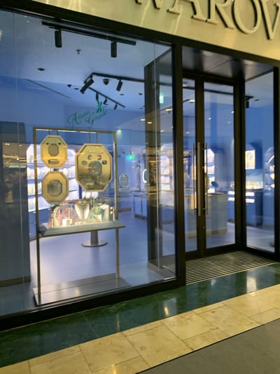 The image shows the facade of a retail store, likely a jewelry or gift shop, as seen through its glass storefront. A sign on the left reads, in part, "Aroma Grune," possibly indicating the store's name. Inside the store, displays of small, gold-colored objects are visible. The store is located in Dublin 16, Ireland. The architectural details suggest a modern shopping center or mall setting. There are no people visible in the image.