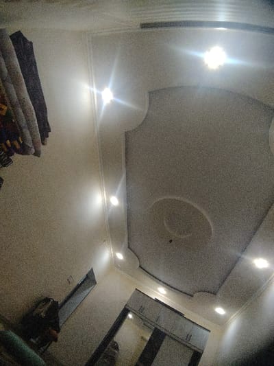 A room's interior is captured from a low angle, focusing on the ceiling. The ceiling features a rectangular design with rounded edges, adorned with recessed lighting. A circular centerpiece is present. Walls are painted in a neutral tone. In the background, a wardrobe with mirror doors is visible. To the left, a partially visible window and hanging clothes add depth.