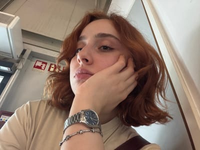 A red-haired person is holding a hand to their face, obscuring part of it. A wristwatch and a bracelet are visible on their wrist. The background suggests an interior space, potentially a vehicle, with a ventilation system and a fire safety sign featuring an extinguisher symbol. The location of the image cannot be determined as Lautzenhausen, Germany.
