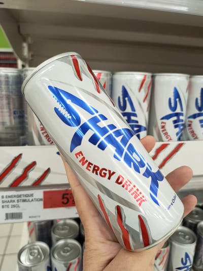 A person's hand holds a white and silver energy drink can with a blue shark graphic and red accents. The text on the can reads "SHARK ENERGY DRINK". In the background, shelves of similar energy drink cans are visible, along with a red price tag displaying "5". The scene appears to be a retail environment, likely a grocery store or convenience store. There is no indication of the location being Cebalat, Tunisia.