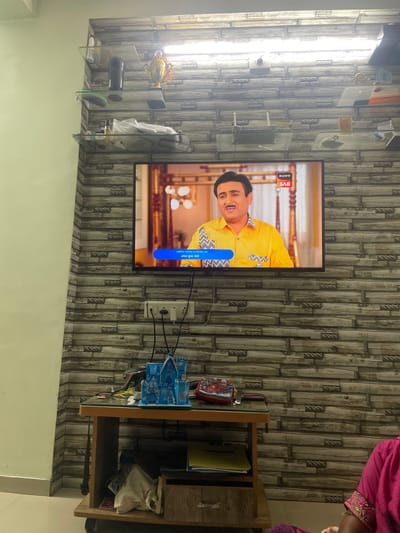 The image shows a living room in Ahmedabad, India, with a television on the wall displaying a scene from a television show. A man in a yellow shirt is on the screen. On a shelf above the television, there are various decorative items. Below the television, there is a wooden table with a blue toy castle and other items. The wall has a textured wallpaper design. A person wearing a pink garment is partially visible in the bottom right corner of the image.