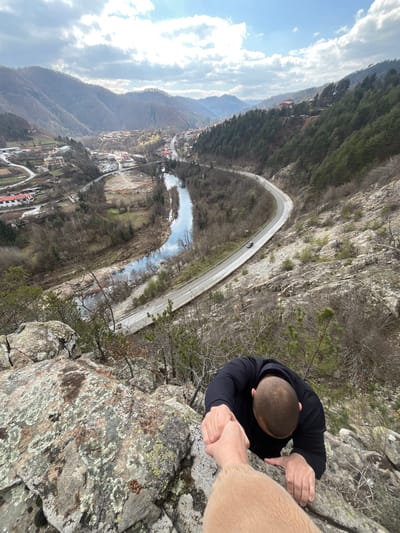 A high-angle view captures two individuals on a rocky incline overlooking a valley identified as Srednogortsi, Bulgaria. In the foreground, a person's light brown-sleeved arm and hand extend downward, gripping the right hand of another person. This second individual, a bald male wearing a black long-sleeved top, is seen from the chest up, leaning forward and using his left hand to grip the moss-covered rock surface. He appears to be ascending the rock face. The immediate foreground consists of rugged, lichen-covered rocks and sparse, dry vegetation.
Below the climbers, a winding paved road, with a visible dark-colored vehicle, parallels a reflective river that meanders through the valley floor. On the far side of the river and road, various buildings, including multi-story structures and houses, are scattered within the settlement of Srednogortsi, extending up the lower slopes of the surrounding mountains. The landscape beyond the town is characterized by forested hills and mountains under a partly cloudy sky, with sunlight breaking through in areas.