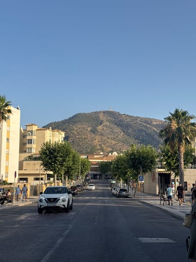 The image captures a street scene in Pla Parcial Platja, Spain, with a prominent mountain in the background. A white Nissan Juke is driving towards the viewer in the foreground, its license plate reading "4978 NKS." Several other vehicles are parked along the right side of the street, and a row of trees lines the sidewalk. On the left side, there are modern buildings, including a tall, cream-colored structure with yellow accents. People are visible walking on the sidewalk and near parked cars. The sky is clear and blue, suggesting fair weather. The mountain in the background is arid and sparsely vegetated, with some telecommunication towers visible at its summit. Palm trees are scattered throughout the scene, characteristic of a Mediterranean coastal town.