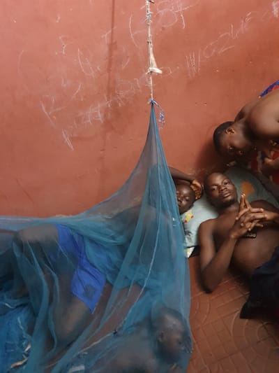 The image captures a group of individuals, appearing to be children or young adults, resting on a reddish-brown floor indoors. At least five individuals are visible, with three clearly in the upper left section, and two more partially obscured by a blue mosquito net in the lower right.
In the upper part of the frame, three individuals lie close together on a light-colored mat or blanket. Their faces are calm, and they appear to be either sleeping or resting. One individual in the center has their hands clasped. A fourth individual's head is partially visible in the top left corner.
The lower right section of the image is dominated by a bright blue mosquito net, stretched diagonally across the frame and tied to a point on a wall or post on the left. Beneath the net, the body of an individual, wearing blue shorts, is visible, suggesting they are also resting. The use of a mosquito net indicates a measure against mosquito-borne diseases, common in regions like Idah, Nigeria.
The floor is predominantly a reddish-brown hue, possibly concrete or a similar material. In the top right corner, a section of the floor appears to be tiled with a brown patterned design. Scattered across the reddish-brown floor are numerous abstract white chalk markings or scribbles. These markings do not form legible words or distinct images but rather appear as random doodles. The lighting is subdued, suggesting an indoor setting perhaps during evening hours or a relaxed moment of the day. The overall atmosphere conveyed is one of quiet rest.