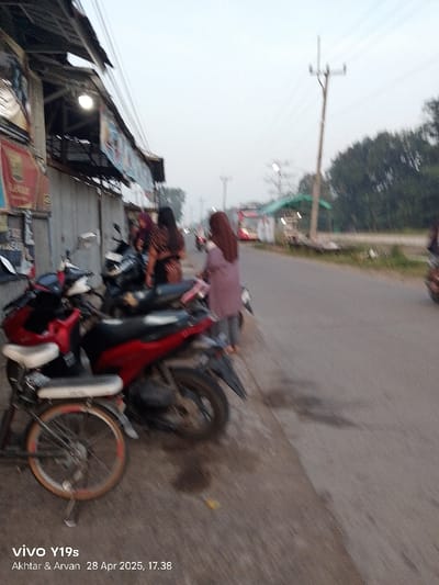 The image shows a street scene in Kecamatan Kroya, Indonesia (latitude: -6.422, longitude: 107.996). Several motorbikes are parked on the side of the street, with one red and black motorcycle in the foreground. Three women are standing in the middle of the frame, looking in different directions. One woman in a dark gray hijab is facing the camera, while the other two are looking away. The street is paved with asphalt and a metal building is seen on the left side of the frame. In the background, there are power lines, trees, and a red bus. It is a cloudy day. The image was taken by a mobile phone.