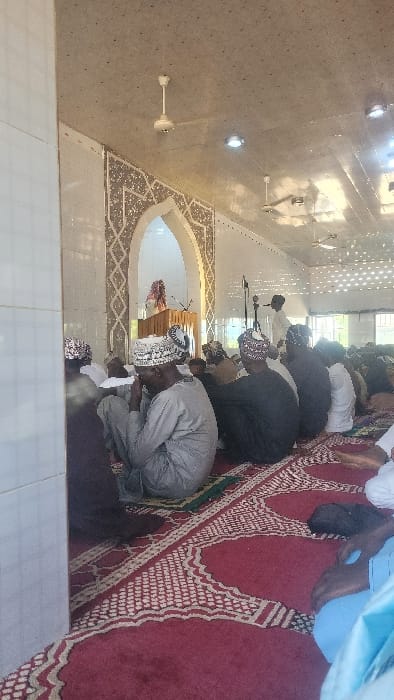 The image depicts an indoor religious gathering within a mosque in Tudun Nupawa, Nigeria. Approximately two dozen men are seated on a red patterned prayer rug covering the floor, arranged in rows facing forward. Most men wear traditional attire, including long robes and patterned skullcaps or kufi hats. In the immediate foreground, one man in a light grey robe and a patterned white cap has his head bowed, with his hands near his face.
At the front of the room, positioned within an ornate arched alcove featuring geometric patterns in white and brown, a figure is standing behind a wooden lectern. This individual, partially obscured, wears a patterned head covering and a light-colored garment, appearing to address the congregation. To the right of the archway, another man stands with a microphone on a stand.
The room features white tiled walls along the left side and a light-colored ceiling equipped with multiple white ceiling fans and recessed light fixtures. Bright ambient light illuminates the scene, with windows visible on the far right allowing natural light. The overall environment suggests a structured religious service or lecture.