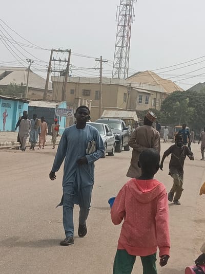 The scene depicts a daylight street view in Hotoro South, Nigeria, characterized by moderate pedestrian and light vehicular activity. The sky is hazy and light-colored.
In the immediate foreground, a dark-skinned adult male wearing a light blue traditional garment (agbada or kaftan) walks towards the left, looking slightly towards the camera. He carries papers or a thin book under his left arm and wears dark sandals. To his right and closer to the viewer, a young, dark-skinned child, seen from behind, walks towards the right. This child wears a pink hooded top with faint white patterns and green trousers, and holds a blue bucket in their left hand.
Behind the foreground figures, a dark-skinned adult male wearing a light brown traditional outfit and a patterned cap walks away from the viewer, angled slightly right. Further back and to his right, a dark-skinned young boy in a torn brown top and trousers runs towards the right, looking back over his left shoulder.
In the mid-ground, two vehicles are visible: a light-colored sedan followed by a dark SUV, both positioned on the right side of the road. On the left side of the street, a group of approximately five dark-skinned adult males, dressed in various light-colored traditional garments, walk together towards the right. One individual in this group wears a red cap.
The environment features an unpaved or lightly paved road surface. Buildings line both sides of the street; one prominent structure on the left has a blue facade with "INTERNATIONAL BOWL" and a boxing graphic painted on it. Various other single and multi-story buildings with flat or corrugated metal roofs are visible. Numerous utility poles with overhead electrical wires crisscross the frame. A tall, red and white lattice telecommunications tower stands prominently in the background on the right. Sparse trees and distant structures complete the horizon.