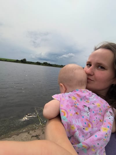 A woman is holding a baby near a body of water. The woman is on the right side of the image, looking towards the camera with a slight smile. The baby is facing away from the camera, nestled against the woman's chest. The baby is wearing a pink onesie with a unicorn and rainbow pattern. The water is dark and reflects the sky, which is cloudy. There is a shoreline with some vegetation visible. The location appears to be natural, possibly a lake or river, and the woman and baby are the primary subjects of the photograph. The setting does not specifically indicate the city of Кояново, Russia, as no identifiable landmarks or text are present.