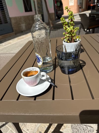 On a light brown slatted table in Andratx, Spain, an outdoor setting is arranged with several items. A small potted plant with green succulents sits near a dark blue drinking glass. A glass bottle of water with a "Dariz" label lies horizontally, close to a small white coffee cup and saucer containing espresso and a spoon. The table appears to be part of a cafe setting. Sunlight casts shadows, with the scene likely captured during daylight. In the background is a building in Andratx, Spain, with exterior walls painted in multiple colors.
