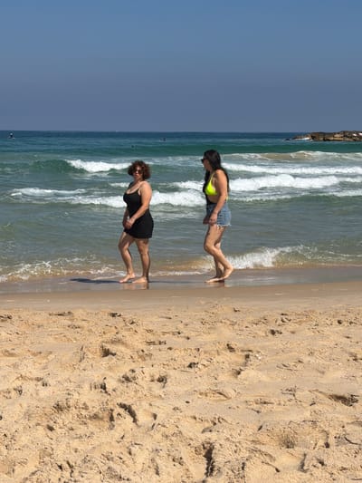 Two women are walking on the beach in Ashkelon, Israel. The woman on the left is wearing a black dress and has curly brown hair. She is looking towards the viewer. The woman on the right is wearing a neon yellow bikini top and denim shorts. She is looking to her left. The ocean is behind them with waves breaking on the shore. The sand in the foreground is light brown and textured. The sky is clear and blue.