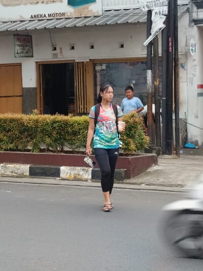 An outdoor scene captured in Cianjur, Indonesia, shows a young female walking on a paved street in the foreground. She is positioned slightly to the left of the frame's center, moving from right to left. She wears a multi-colored short-sleeved top with "13" printed in white on the front, black athletic leggings with a white logo on the left thigh, and brown flat sandals. A purple and black backpack is worn on her shoulders. Her dark hair is pulled back. In her right hand, she holds a clear plastic cup with a white lid, and in her left hand, a folded object, possibly a paper or small electronic device.
Behind her, to her right, a low planter box with a brown concrete edge contains dense green and reddish-leafed bushes, separating the street from a sidewalk area. Beyond this, a white single-story building is visible, identified by a sign on the facade reading "ANEKA MOTOR". To the left of the main entrance, a smaller, older-looking sign also reads "ANEKA MOTOR" with additional text below, including "CIANJUR". The building has a corrugated roof, two rectangular window openings, and an open doorway revealing a darker interior where a "GULF" logo is discernible.
Further right, a male subject in a light blue short-sleeved shirt is partially visible behind the bushes, standing near a dark utility pole structure. This utility pole structure has multiple dark poles and wires, with a torn white sign attached to one, showing "TERAPI TINGGI BADAN" text and indistinct figures. Another label on the pole reads "CJR-100-474". A blurred white object, consistent with the front wheel and fender of a moving motorcycle or scooter, occupies the lower right corner of the frame, indicating traffic on the street. The street surface is gray asphalt, with a curb featuring weathered white and black paint markings. The lighting suggests an overcast day.