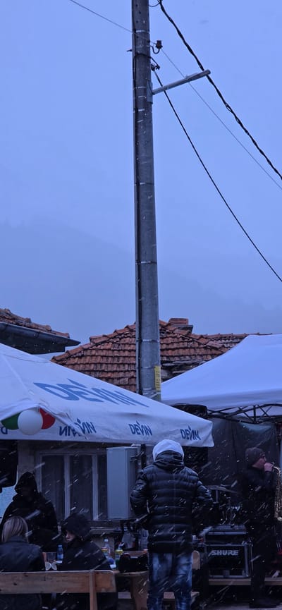 The image depicts a scene in Breze, Bulgaria, during a snowy day. A grey utility pole stands vertically in the left foreground, with electrical wires extending from its top. Behind the pole, there is a building with a red tiled roof. To the right, a white tent with blue "Devin" lettering is partially visible. Under the tent, several people are present; one plays a saxophone. There is a white box near them with a "Shure" logo. Snowflakes are falling throughout the scene.